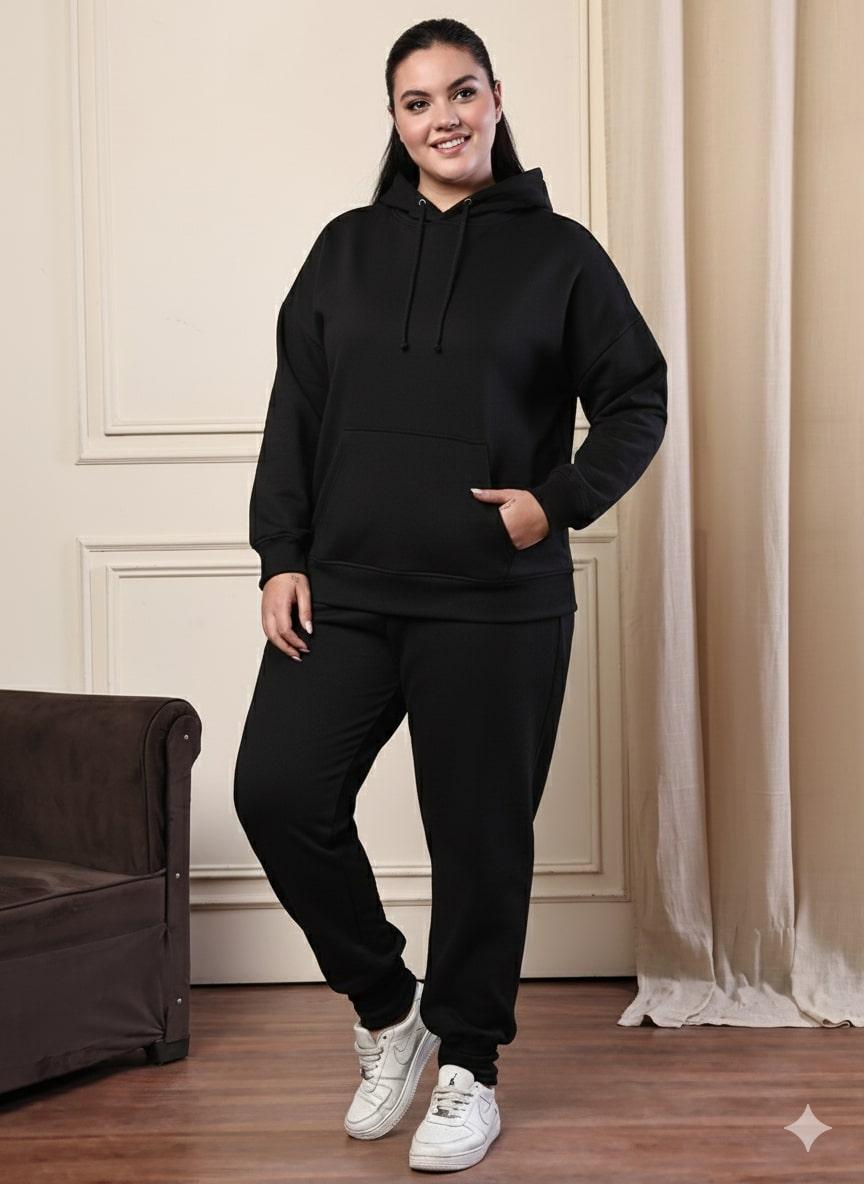 Person wearing a black hoodie and pants standing in a room with a brown couch and white curtains.