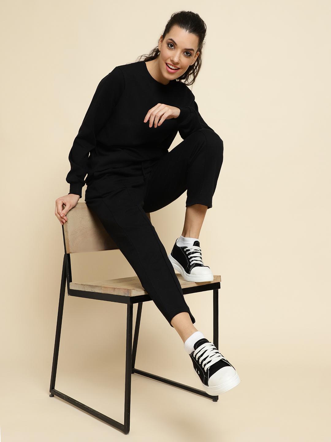 Woman sitting on a chair wearing a black tracksuit with white sneakers against a beige background