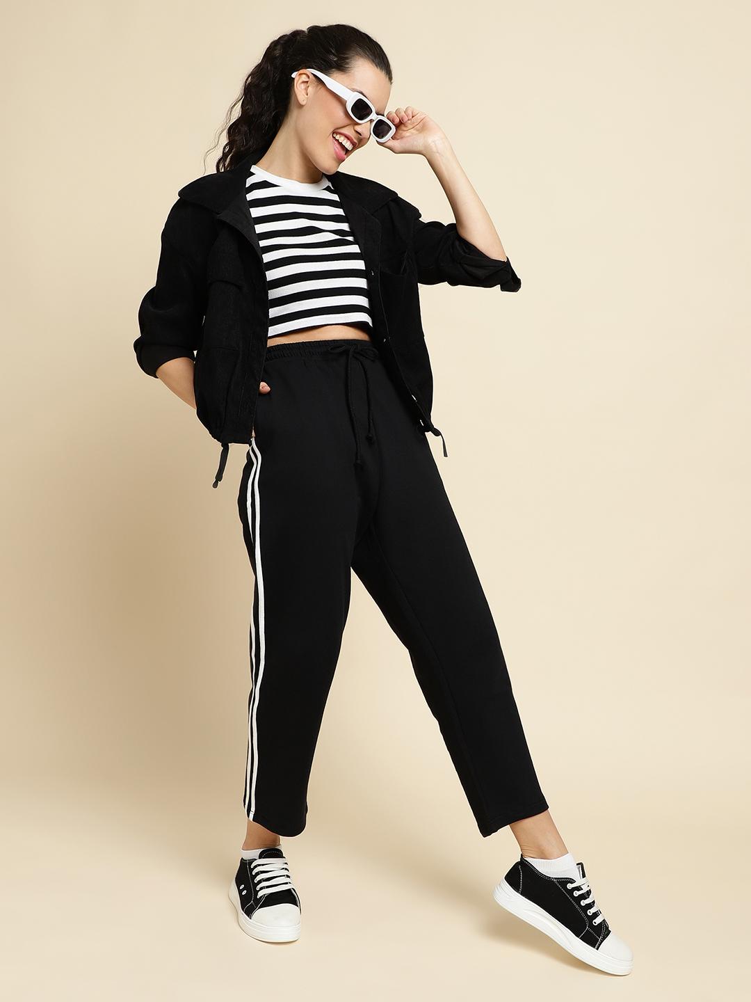 Woman wearing a black jacket, striped shirt, and black track pants with white stripes on a beige background
